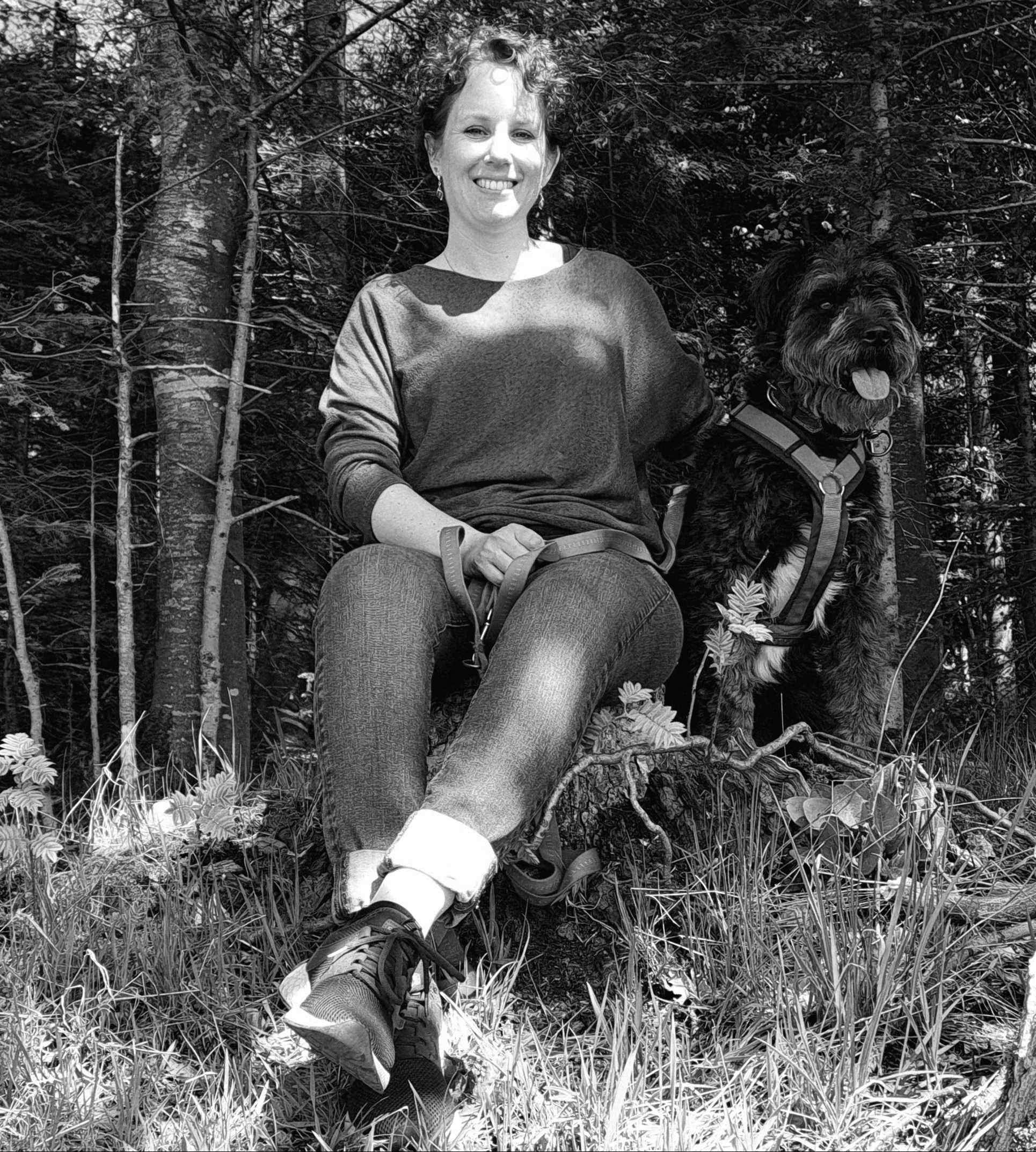 TBlack and white portrait of social anthropologist Ariane Zangger sitting in a lush, wooded environment. She is smiling warmly, seated beside a scruffy, dark-colored dog in a harness, evoking a sense of connection with nature.